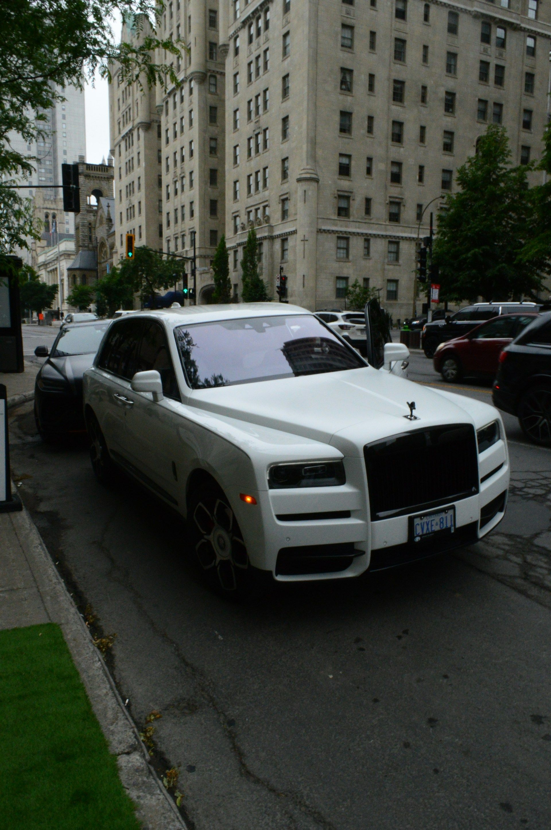 A white Rolls-Royce Cullinan SUV parked on a city street next to a tall, historic stone building.
