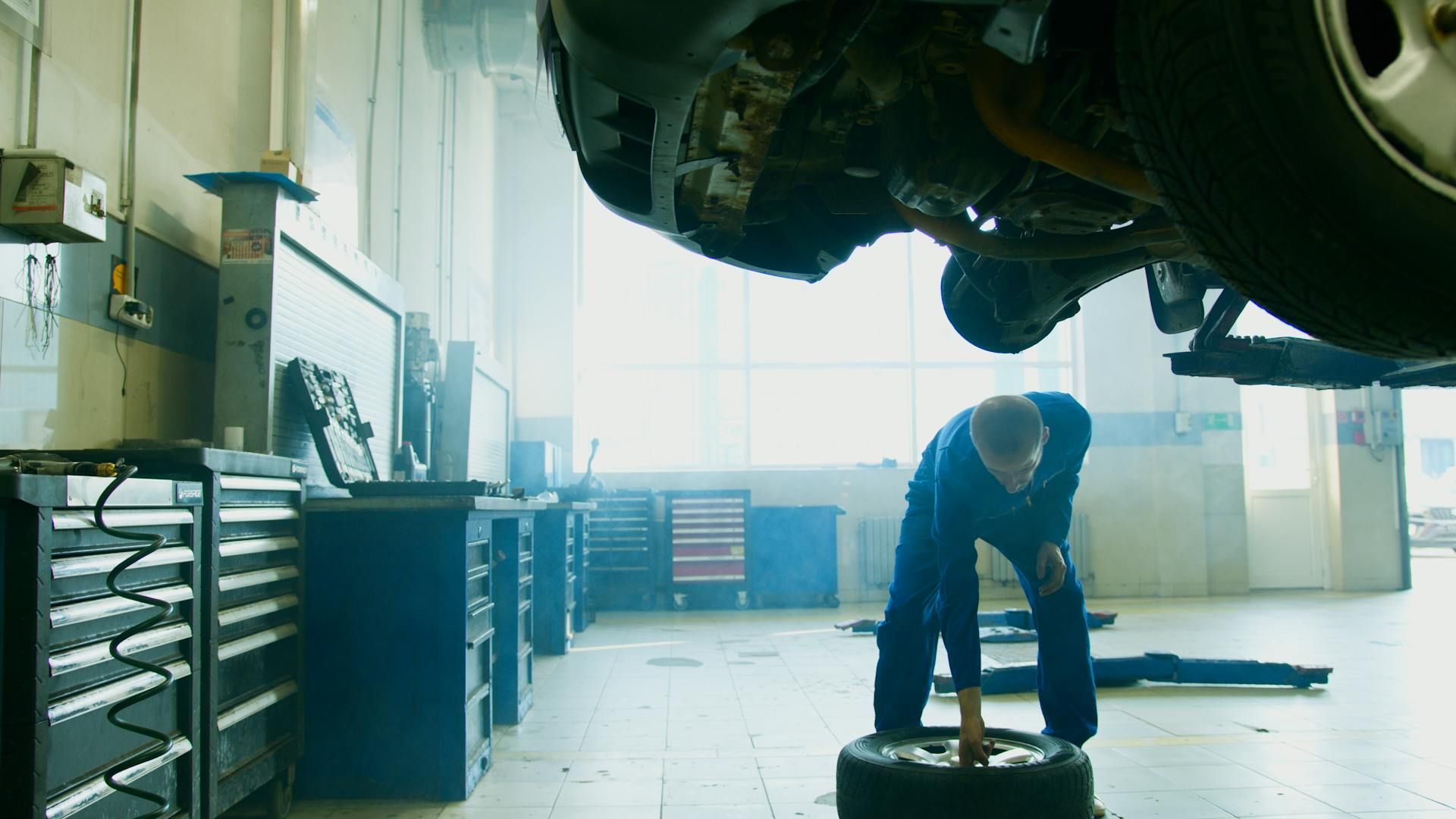 A mechanic in blue workwear inspecting a tire in a bright, modern auto repair shop with a car lifted overhead.