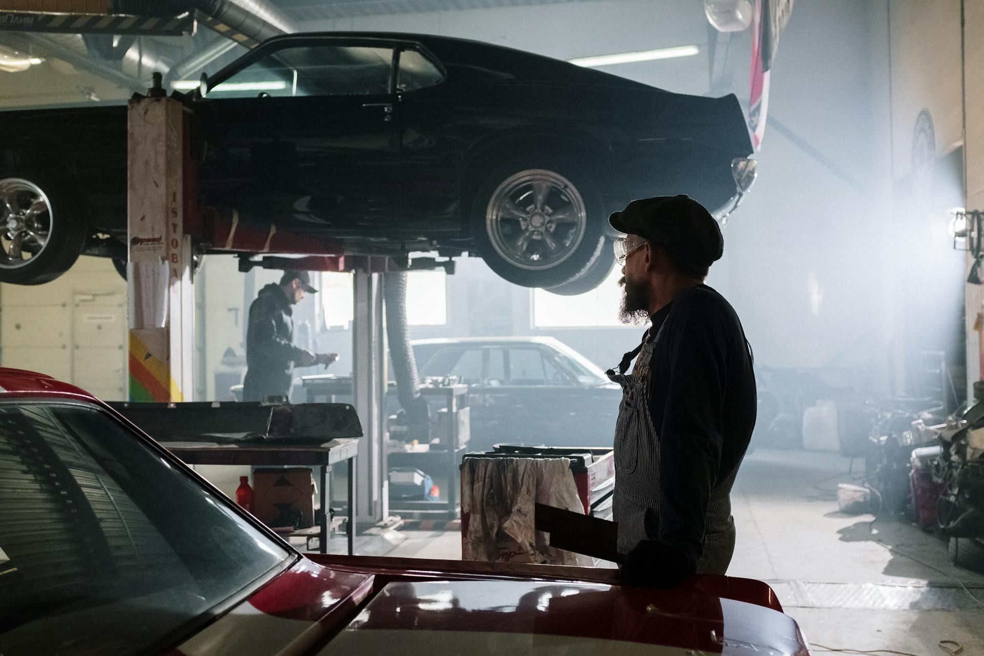 Two mechanics work in a bright, hazy auto repair shop, with a black car lifted on a rack and a red car in the foreground.