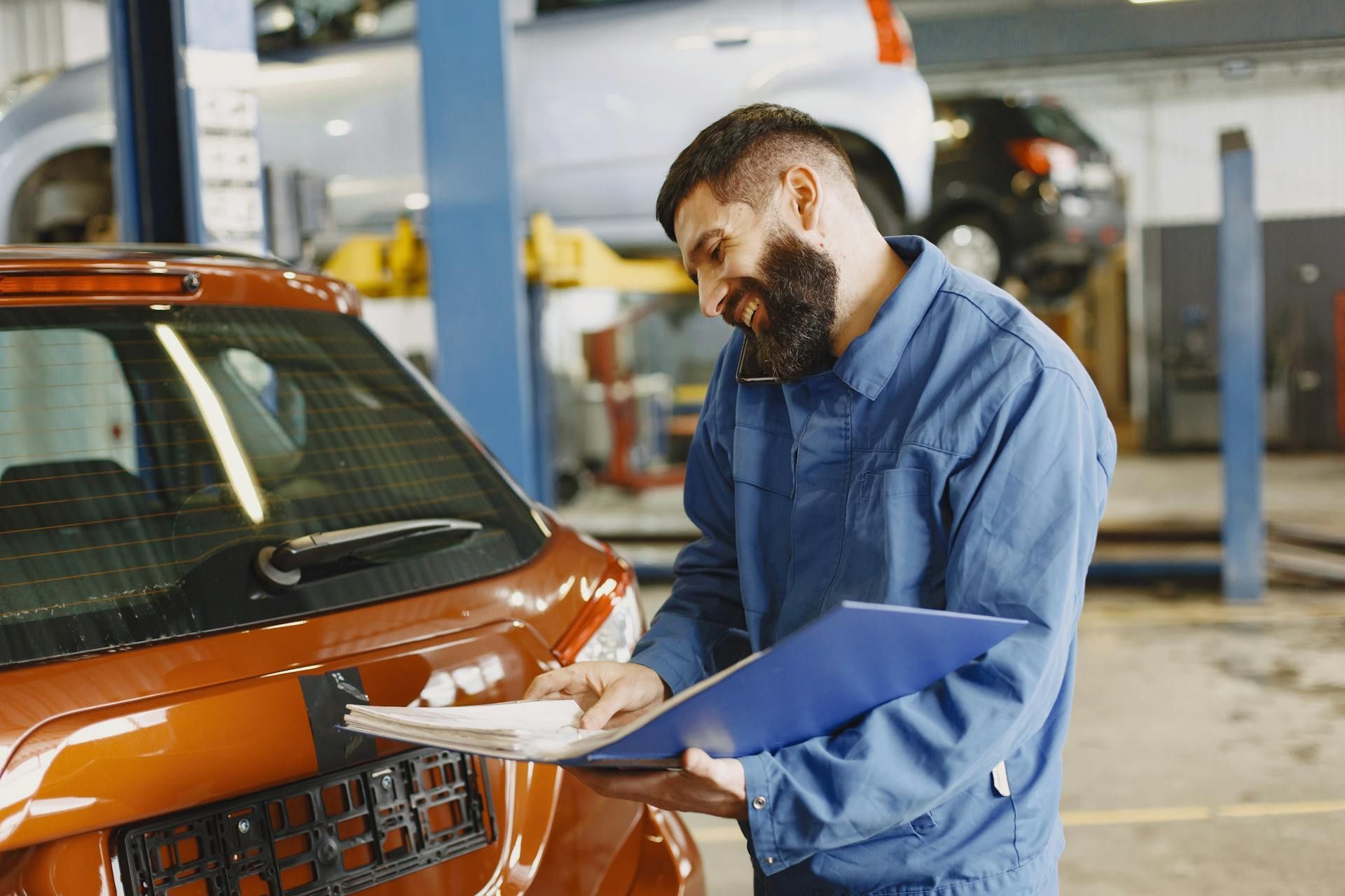 A mechanic in a blue uniform checks a document while standing next to an orange car in an automotive repair shop.
