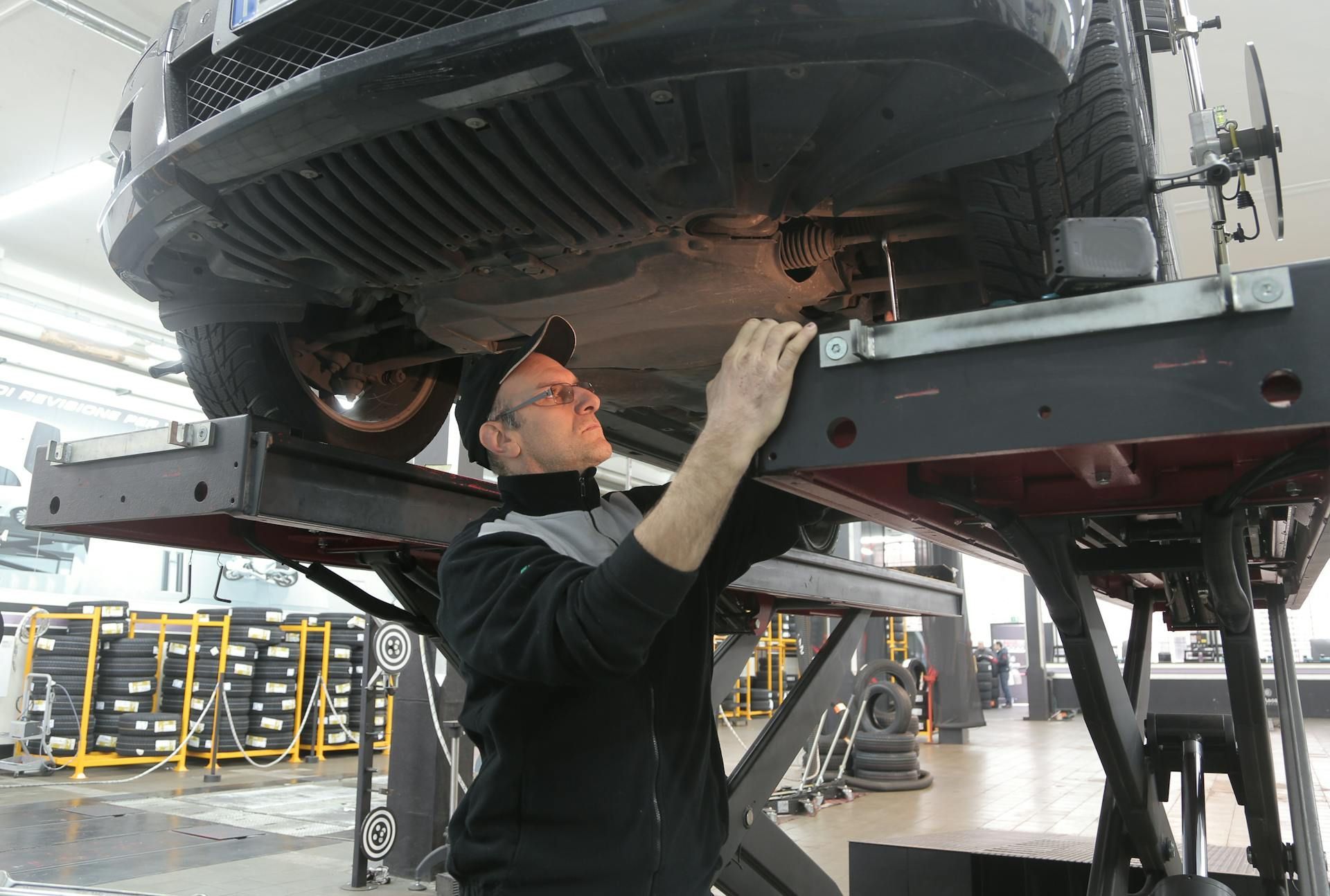 A mechanic in a black jacket inspecting the underside of a car raised on a hydraulic lift inside an auto repair shop.