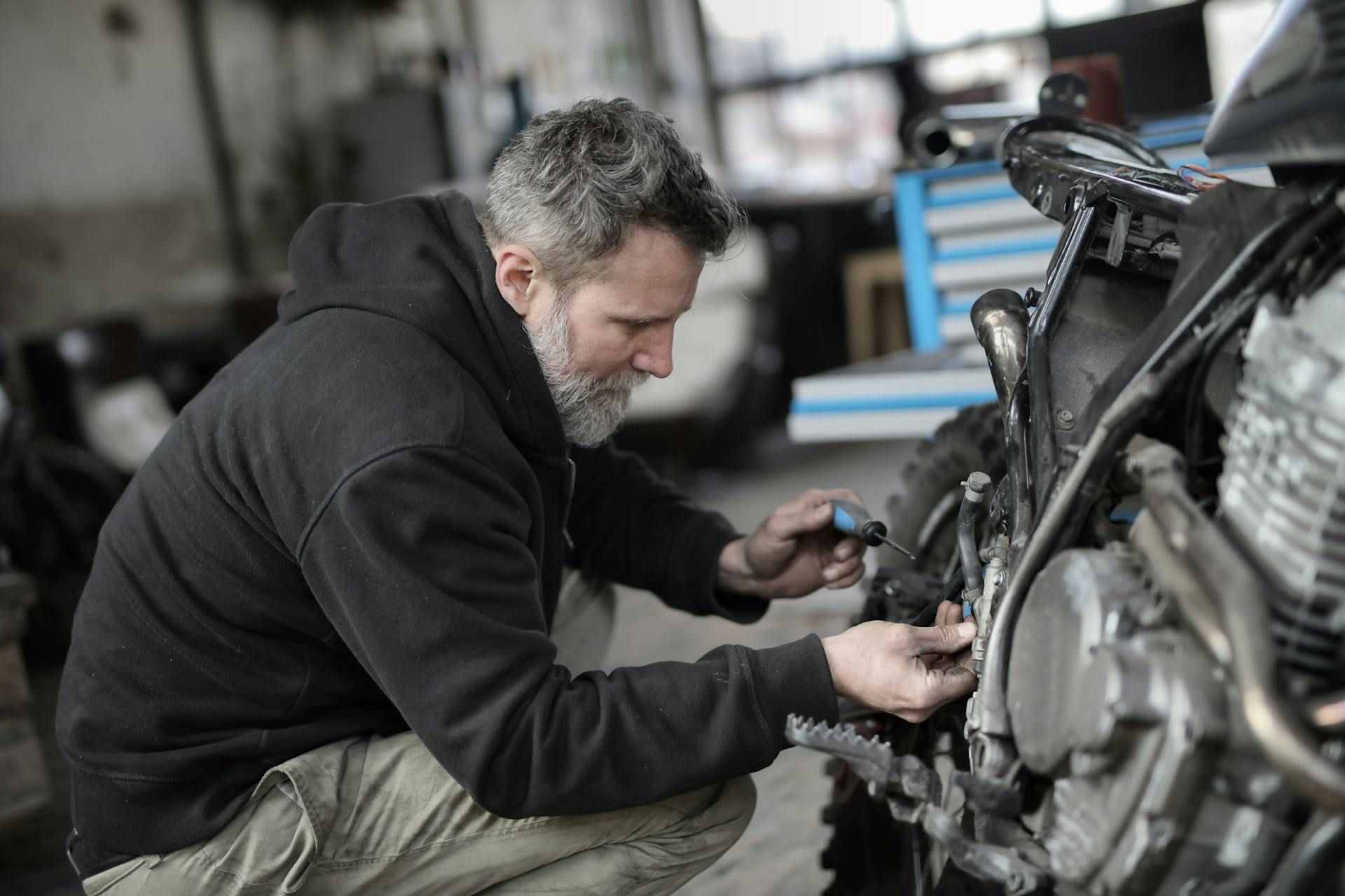 A bearded mechanic in a black hoodie inspects the engine of a motorcycle in a dimly lit garage.