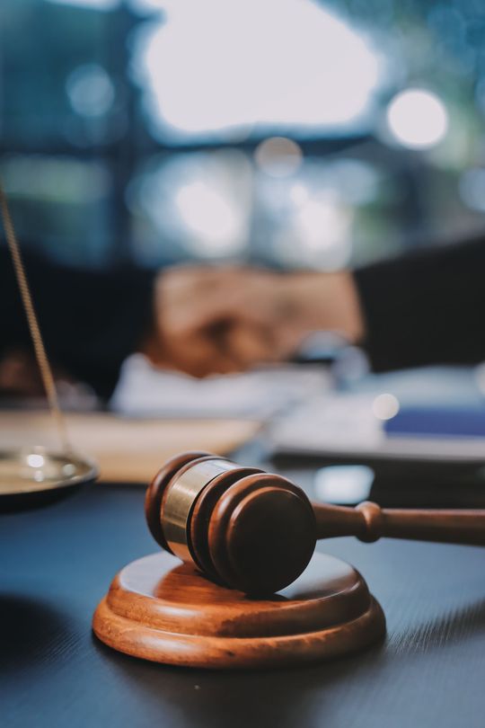 A wooden judge's gavel rests on a matching sounding block on a dark desk, with a blurred courtroom background.