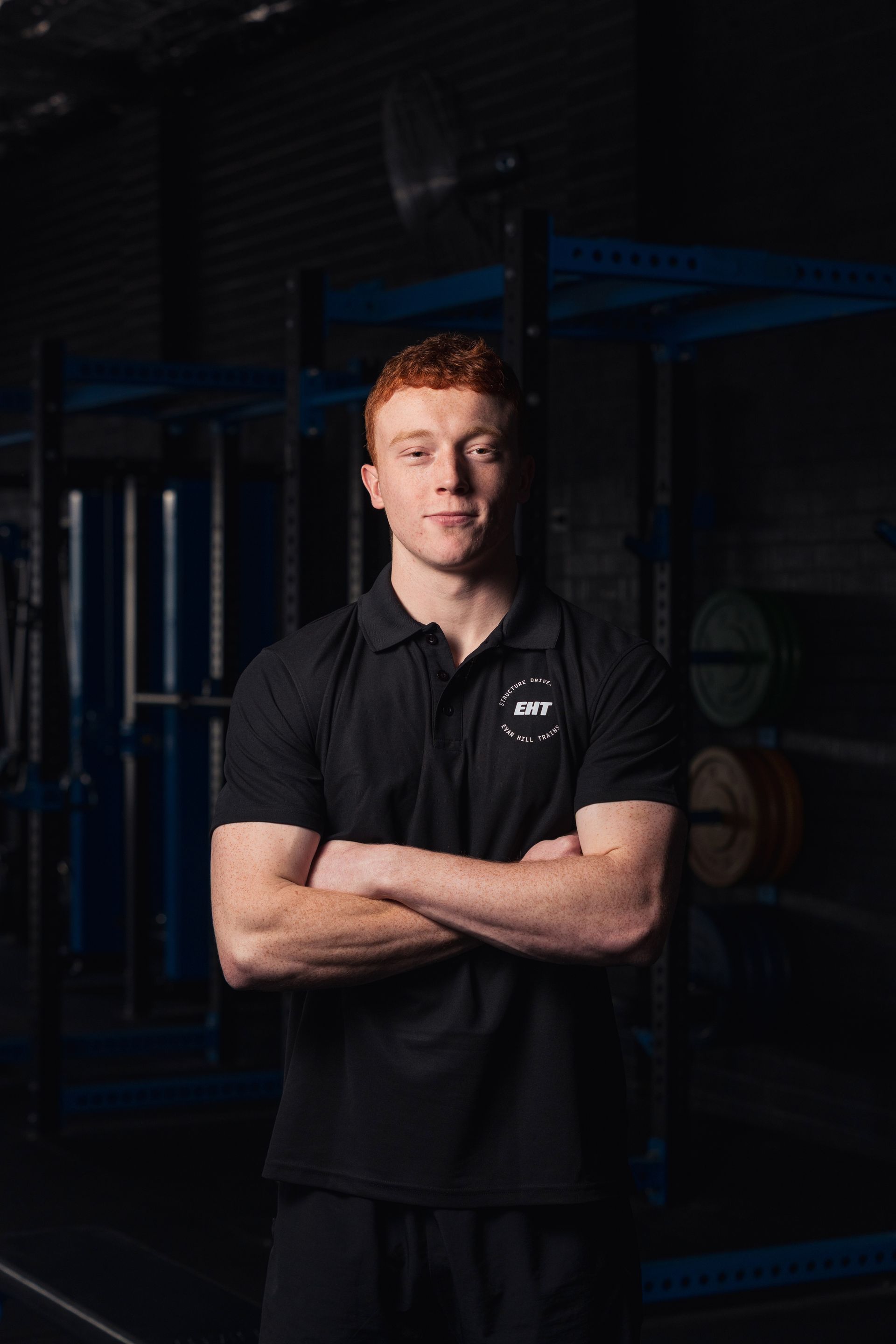 Man standing with arms crossed in a dark gym, wearing a black polo shirt, facing the camera.
