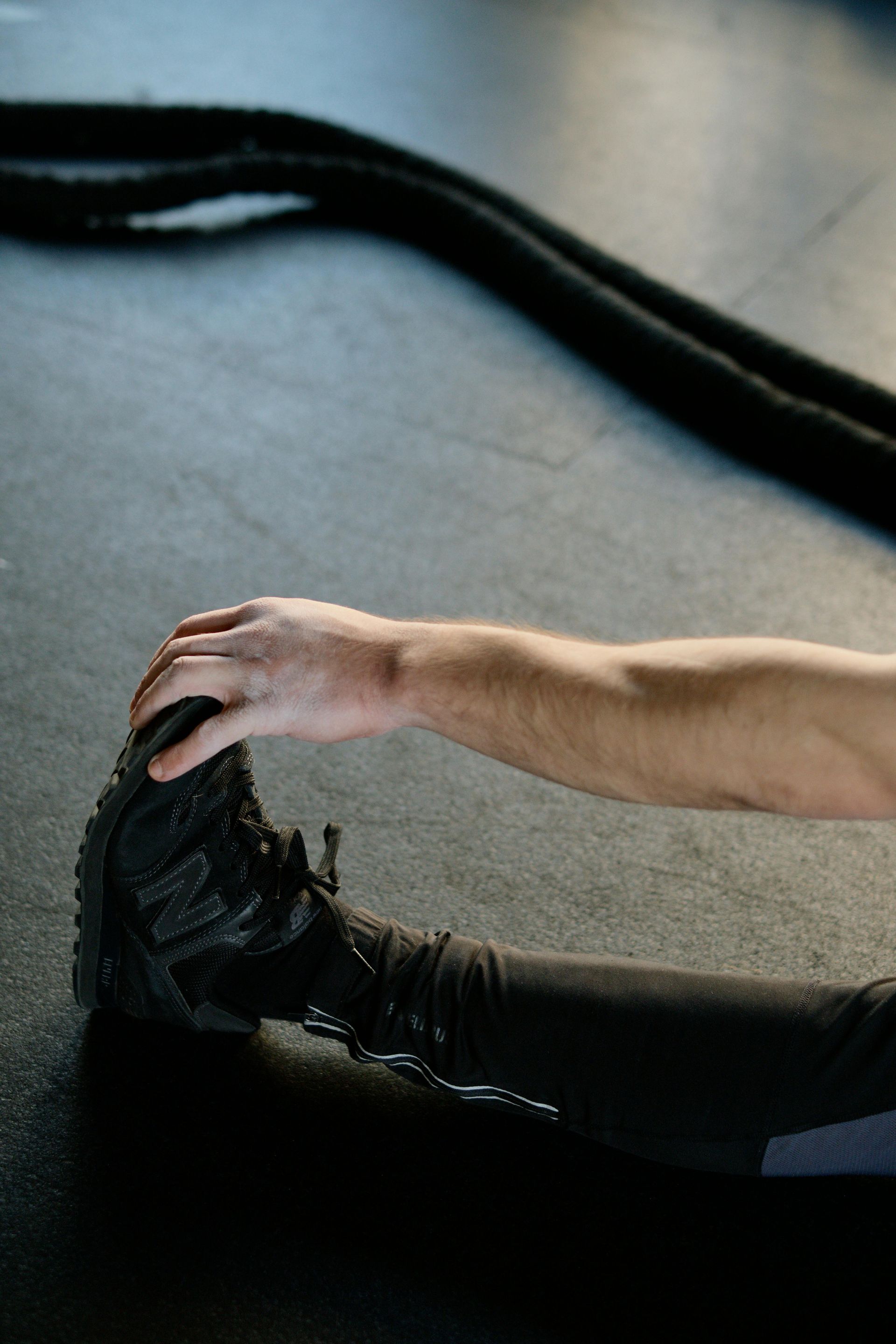 Hand reaching toward a black shoe on a concrete floor, with a thick black rope in the background