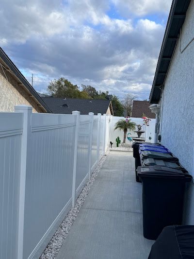 A white fence surrounds a sidewalk with trash cans in front of a house.