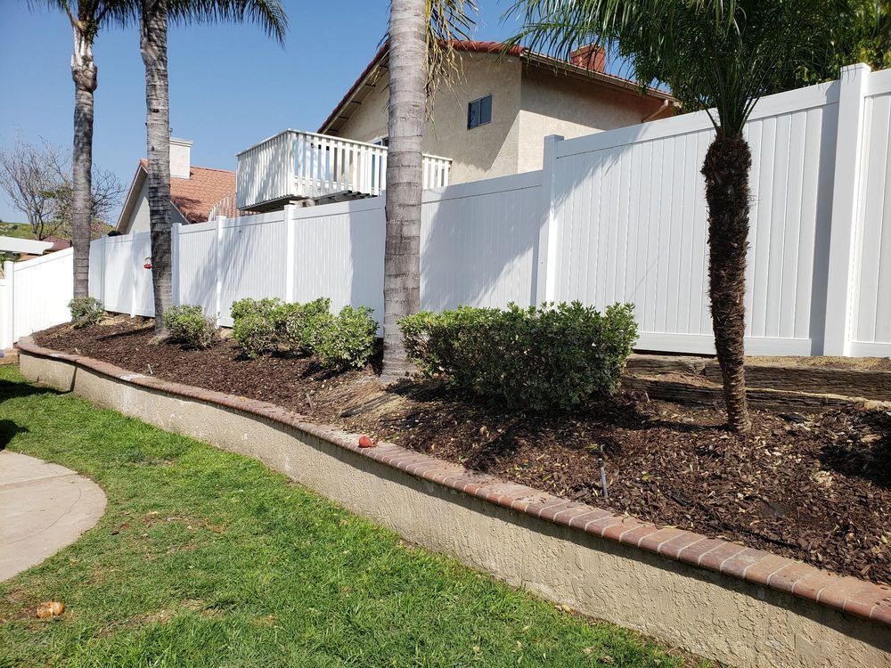 A white fence surrounds a lush green yard with palm trees.