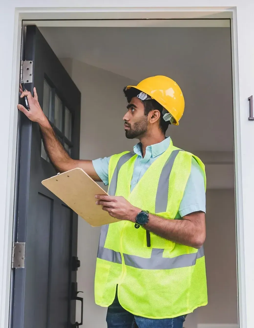 A man wearing a hard hat and safety vest is standing in front of a door holding a clipboard.
