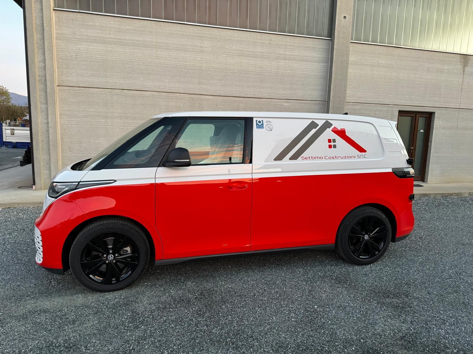 Furgone Volkswagen ID. Buzz rosso e bianco con ruote nere, parcheggiato davanti a un edificio.