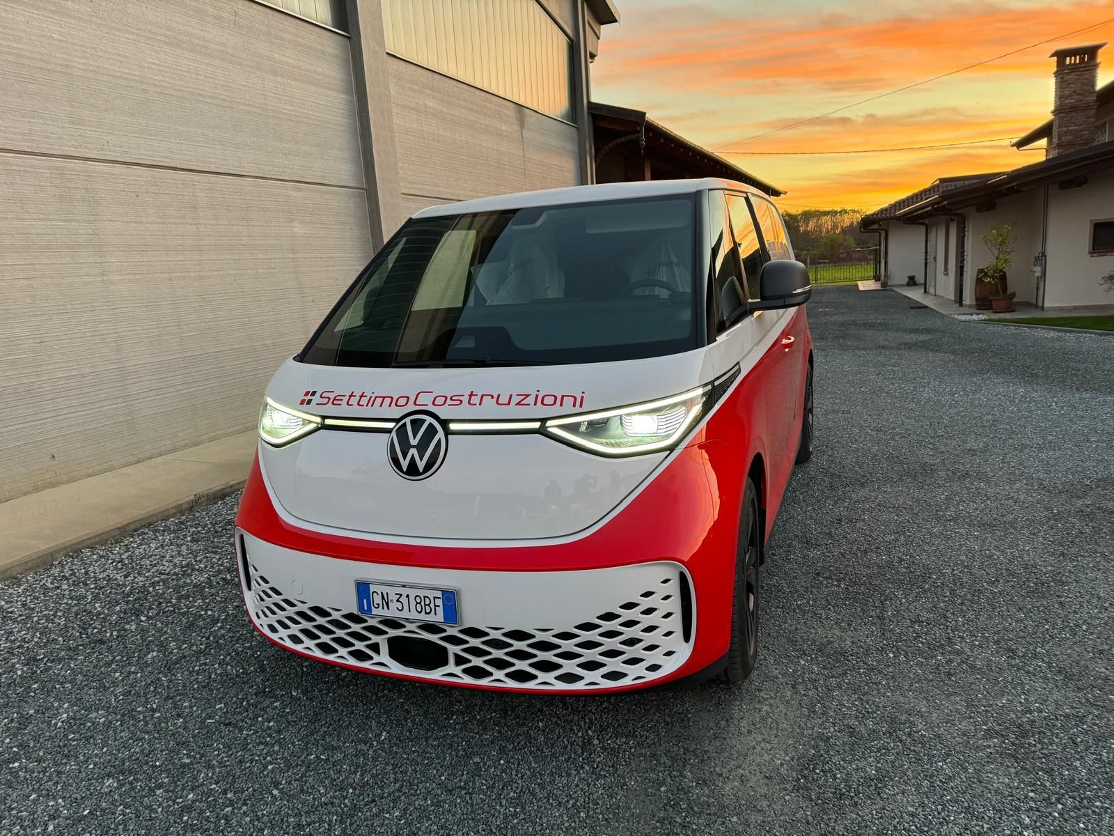 Un furgone Volkswagen ID. Buzz rosso e bianco parcheggiato fuori da un edificio al tramonto.