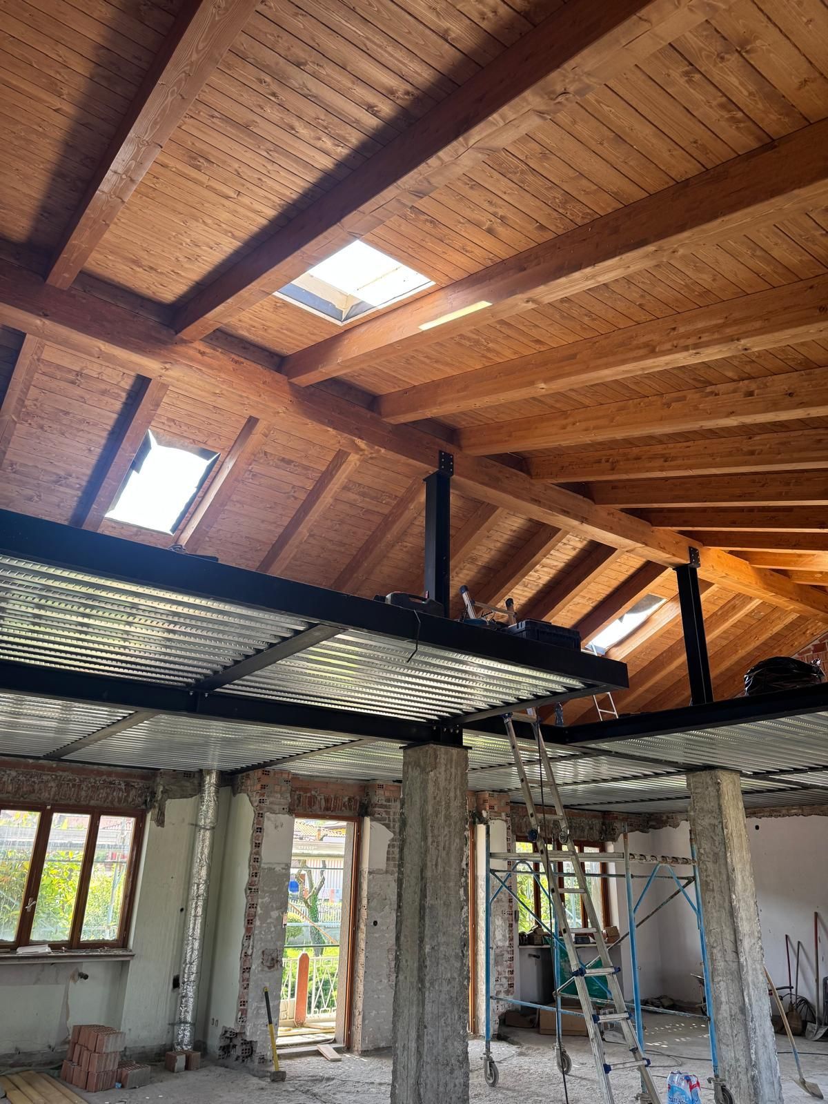 Vista interna di un edificio in fase di ristrutturazione, caratterizzato da soffitto in legno, soppalco in metallo e lucernari.