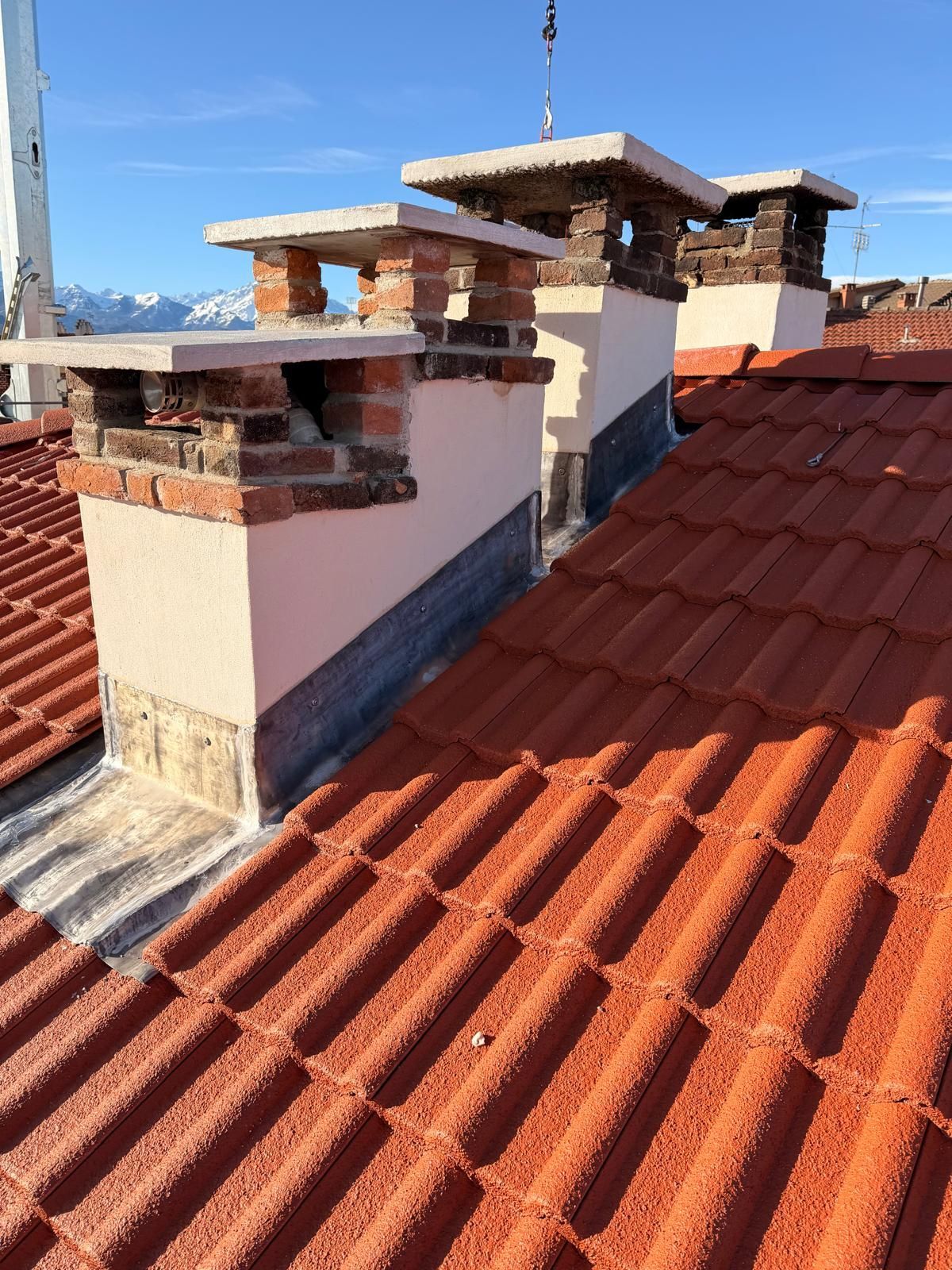 Tetto di tegole rosse con tre camini in mattoni contro un cielo azzurro, sullo sfondo delle montagne.