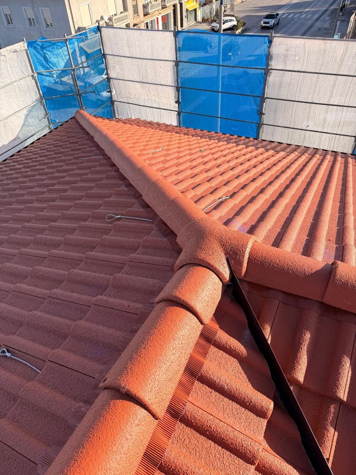 Tetto di tegole rosse con grondaia centrale, visto dall'alto, telo blu ed edifici sullo sfondo.