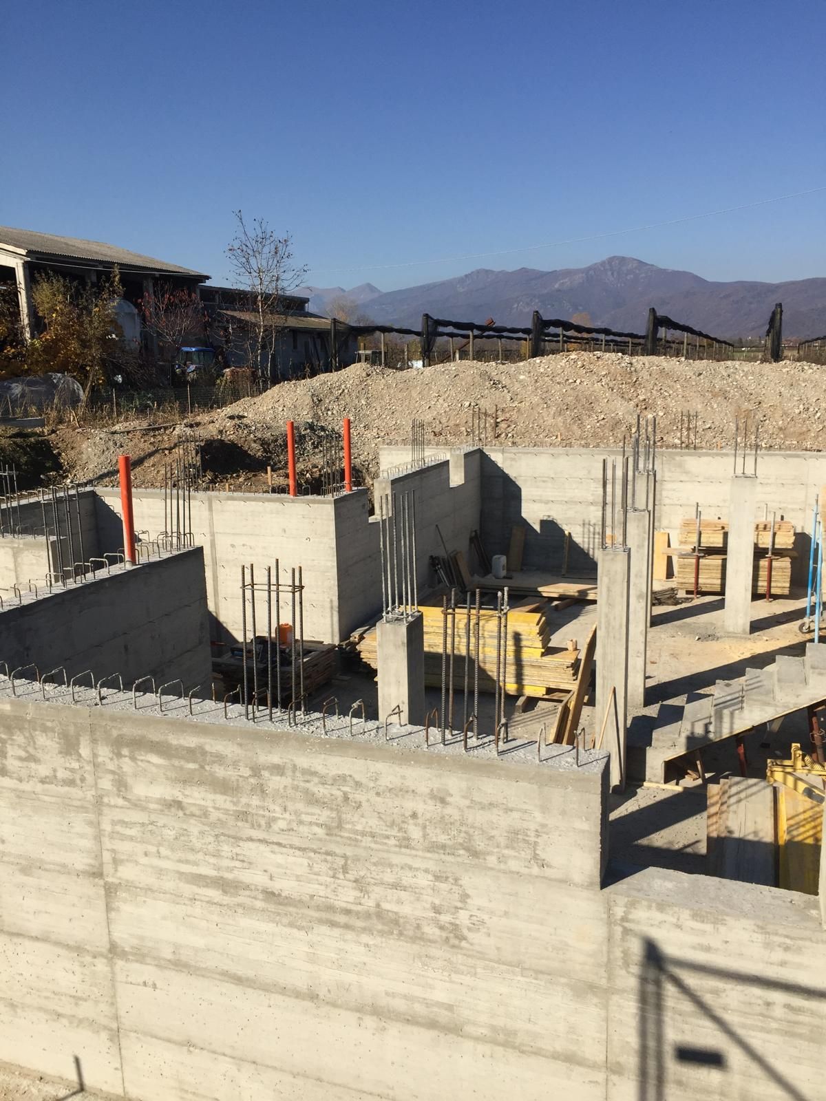 Cantiere con fondamenta in cemento e barre di armatura, sullo sfondo di una collina e di un cielo azzurro.
