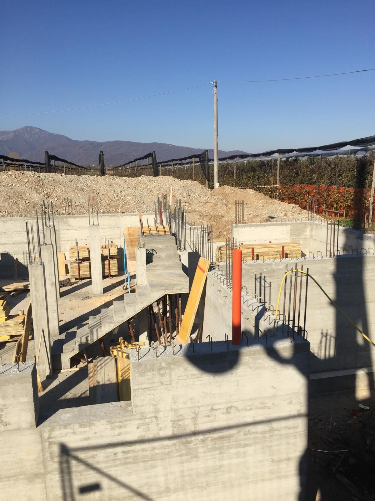 Fondamenta in cemento in costruzione, con barre di armatura, supporti in legno e un mucchio di ghiaia in un luogo soleggiato.