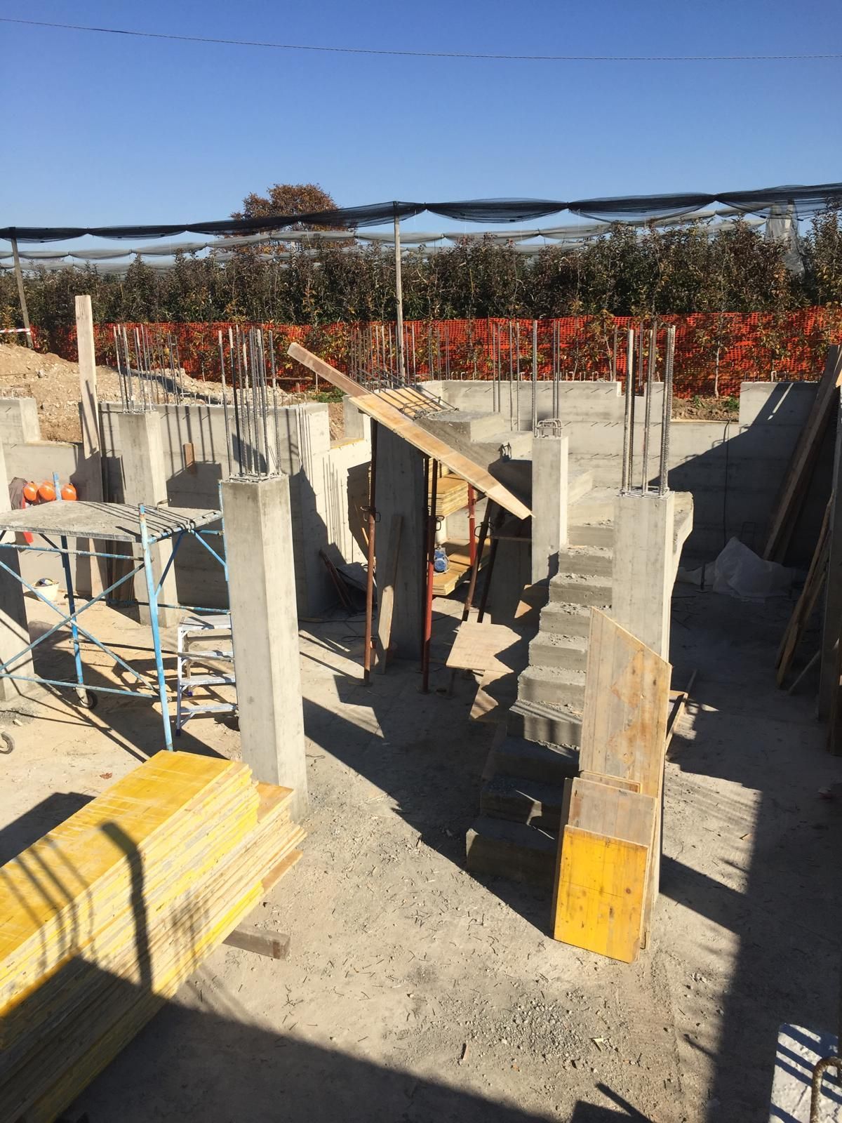 Cantiere: pilastri di cemento, muri e impalcature sotto un cielo azzurro, con assi di legno e reti di sicurezza arancioni visibili.