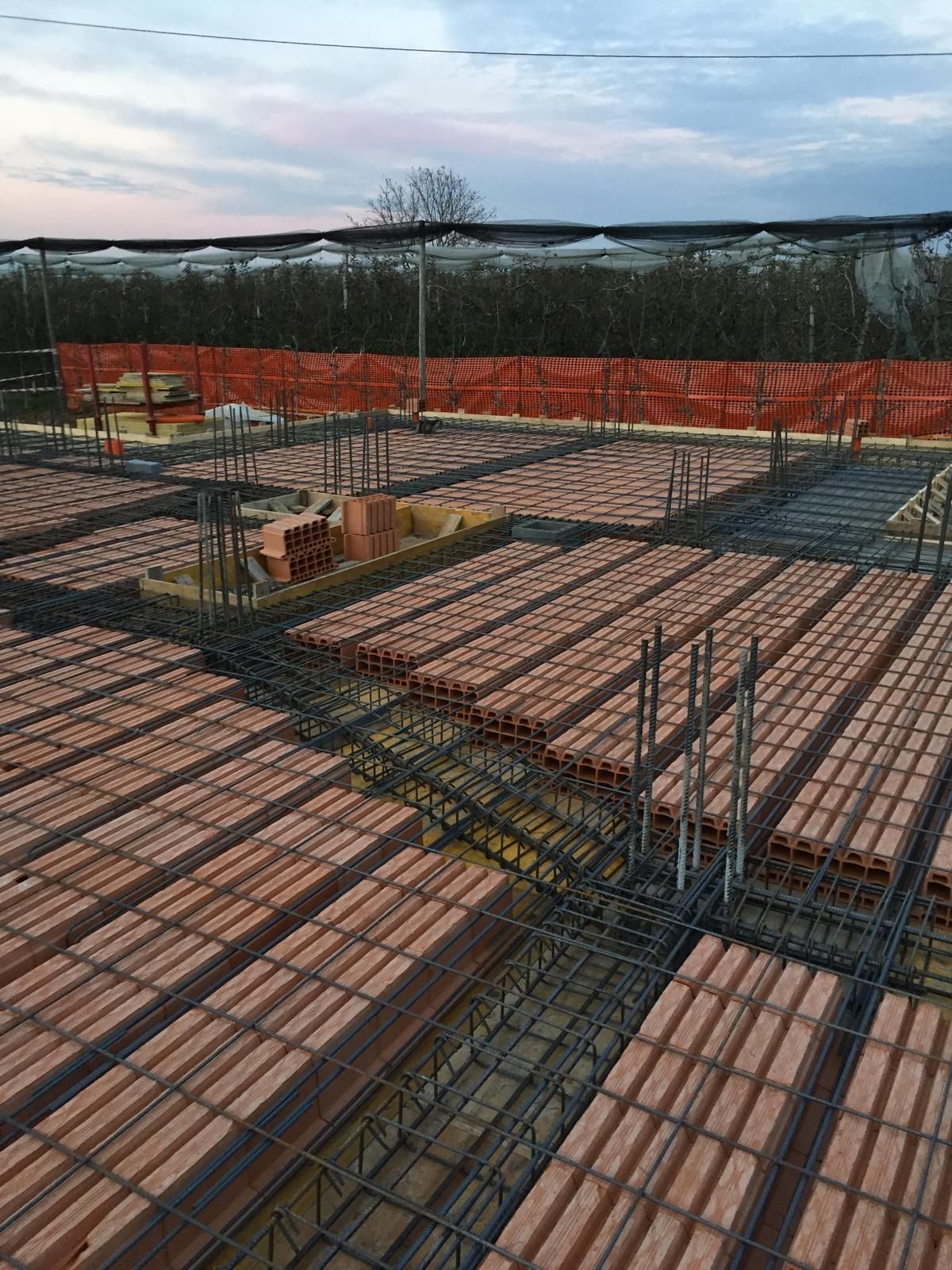 Cantiere con soletta di cemento parzialmente realizzata. Mattoni rossi tra barre d'armatura, recinzione di sicurezza arancione, cielo nuvoloso.