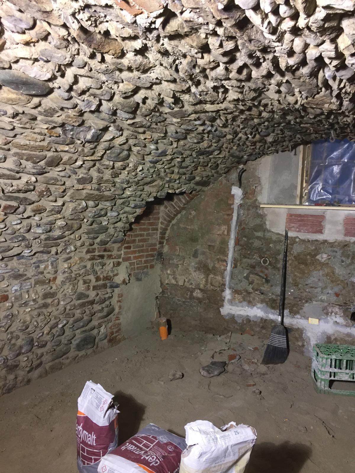 Interno di una cantina con arcate in pietra, sacchi di materiale, una finestra coperta di plastica e una pala.