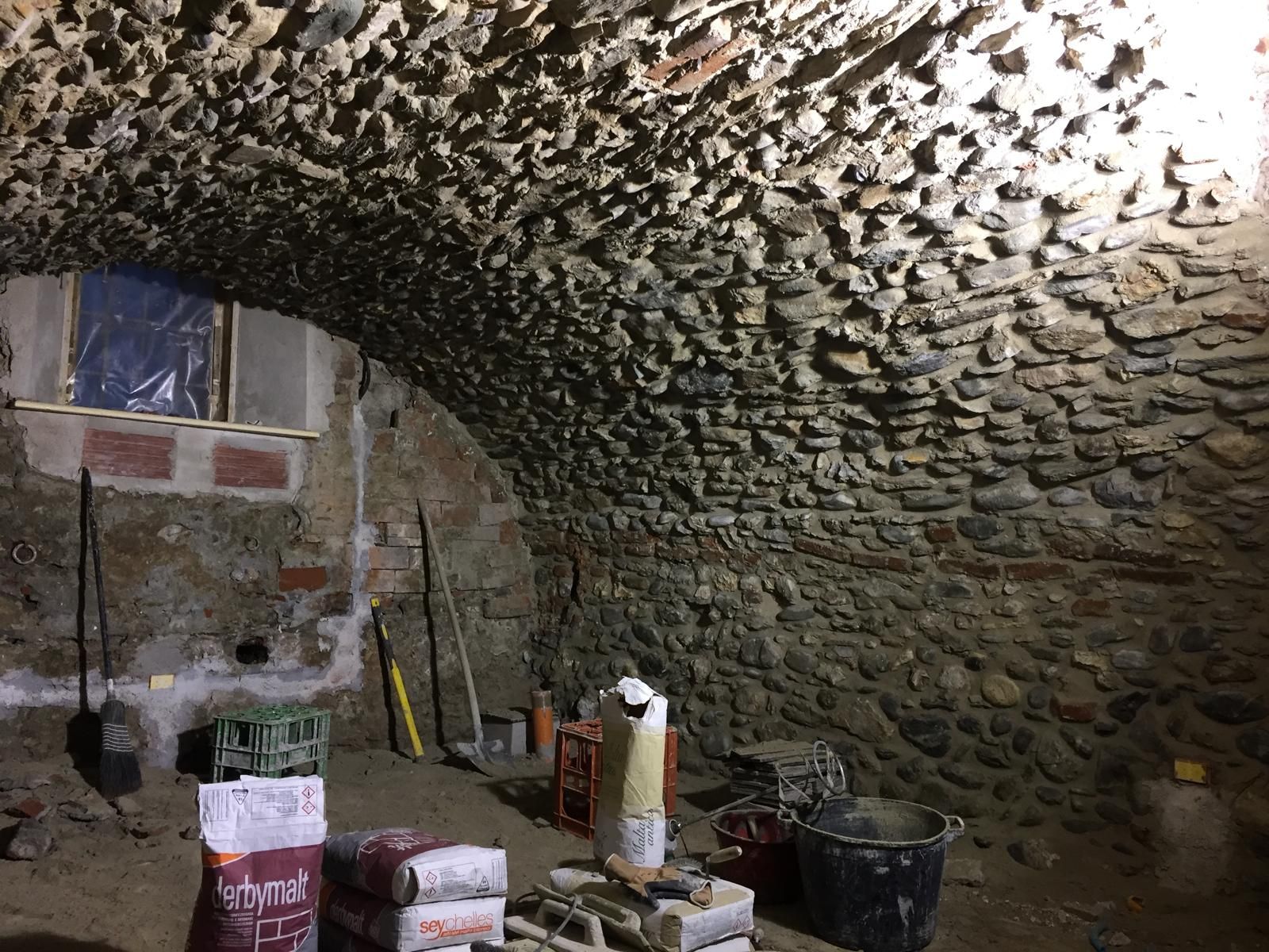 Interno di una cantina con volta in pietra. Attrezzi e materiali da costruzione sono sparsi sul pavimento in terra battuta. Una finestra si apre sul muro di mattoni.