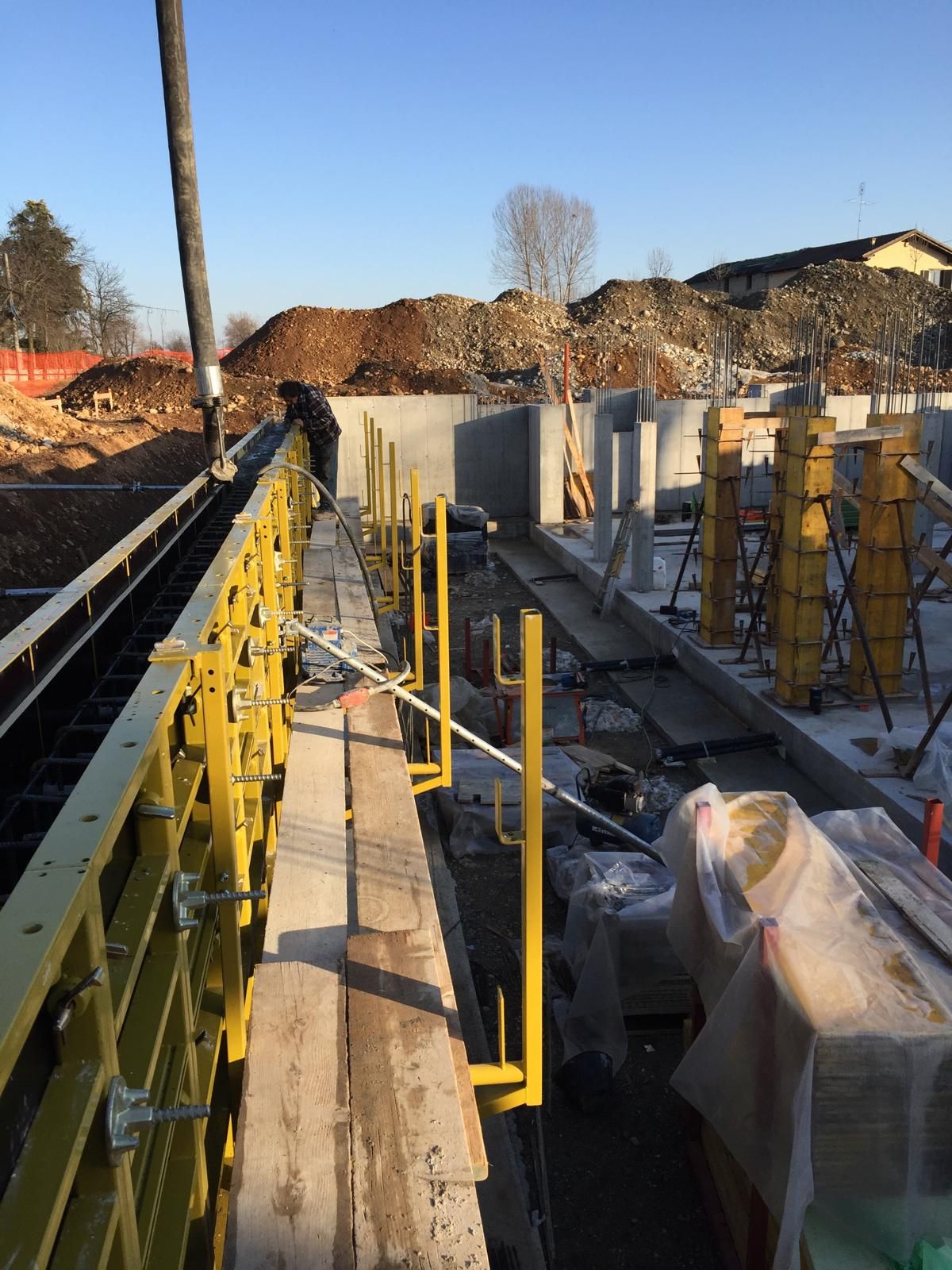 Cantiere con impalcature gialle e casseforme in cemento; sullo sfondo un mucchio di terra.