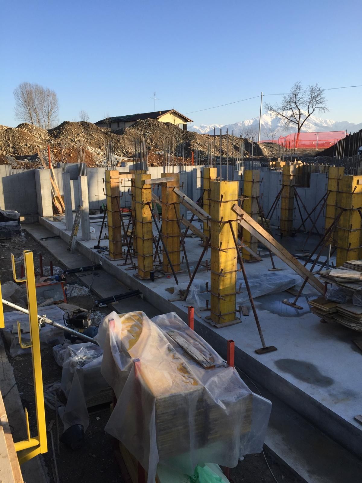 Cantiere con casseforme in cemento giallo per colonne, travi di sostegno e sfondo di macerie e montagne.