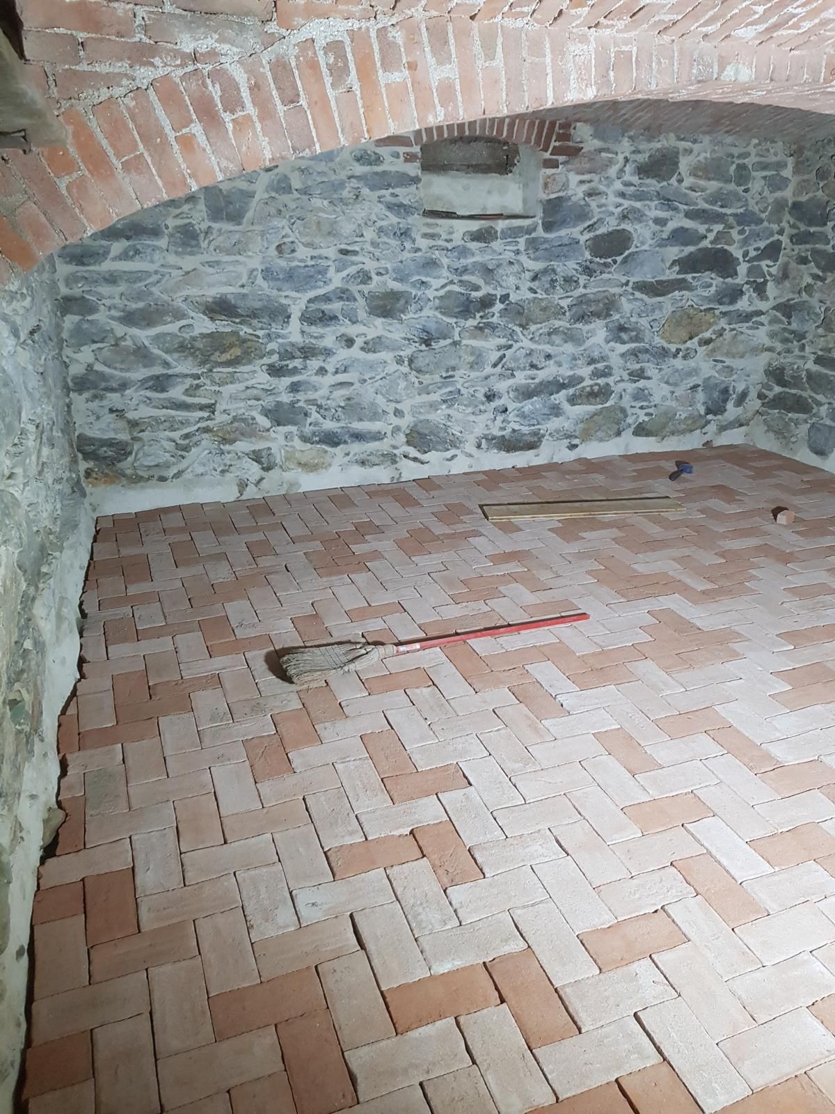 Stanza con pareti in pietra, soffitto a volta in mattoni e pavimento in mattoni, con una scopa e un attrezzo dal manico rosso.