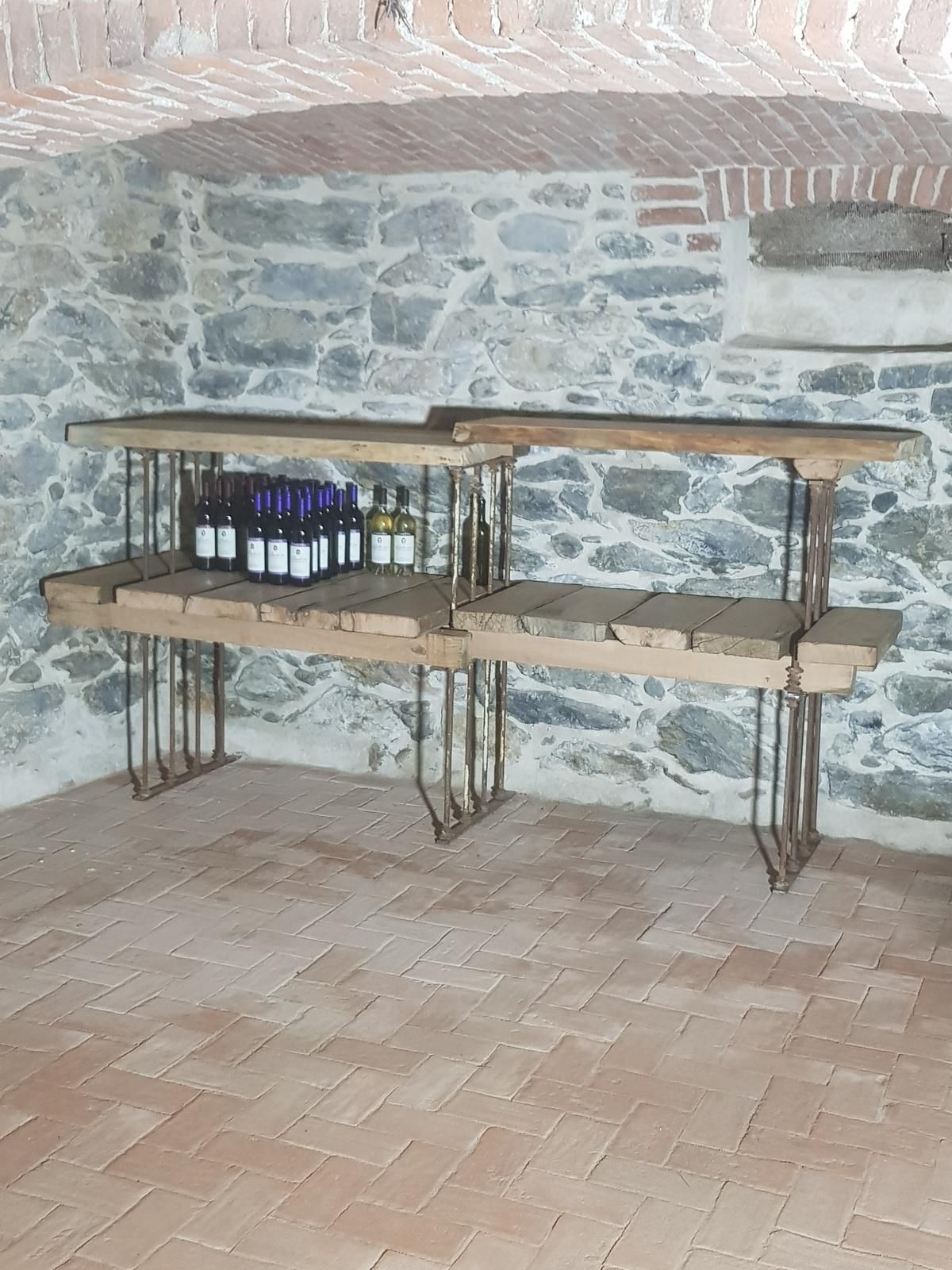 Rustico bancone in legno con gambe in metallo che sorregge bottiglie su uno sfondo di muro in pietra.