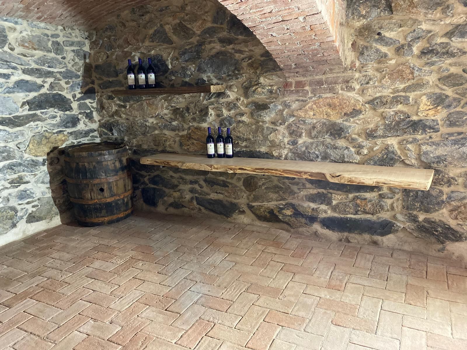 Cantina con pareti in pietra e pavimento in mattoni. Le bottiglie di vino sono appoggiate su scaffali di legno. Nelle vicinanze si trova una vecchia botte.