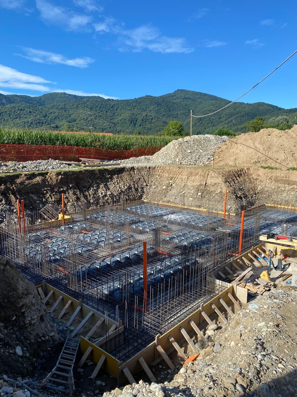 Cantiere con struttura in tondino di ferro all'interno di uno scavo profondo, contesto rurale con montagne sullo sfondo.