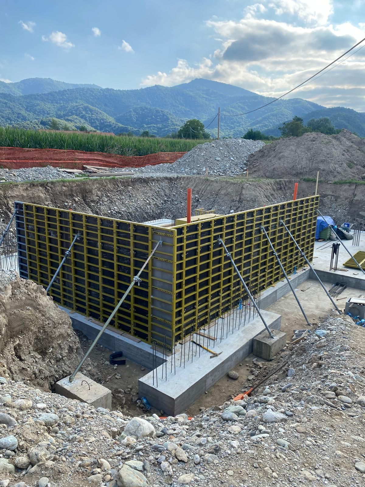 Cantiere con fondamenta in cemento e muri con struttura gialla, con montagne sullo sfondo.