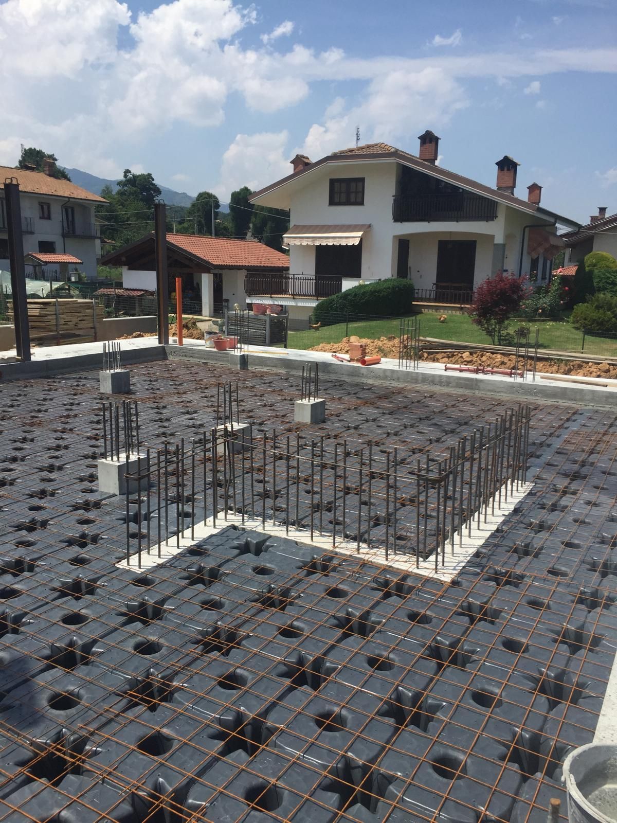 Cantiere con griglia di barre d'armatura su casseforme nere, pronto per il getto di calcestruzzo; case sullo sfondo.