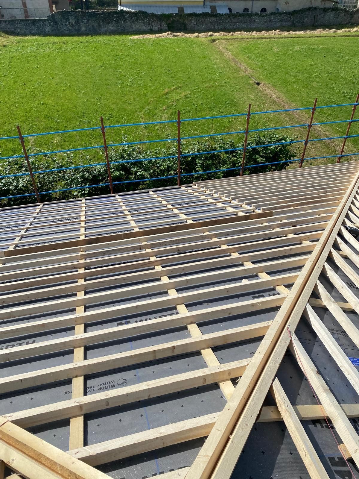 Costruzione del tetto: travi in ​​legno, rivestimento e un sottofondo scuro; campo verde sullo sfondo.