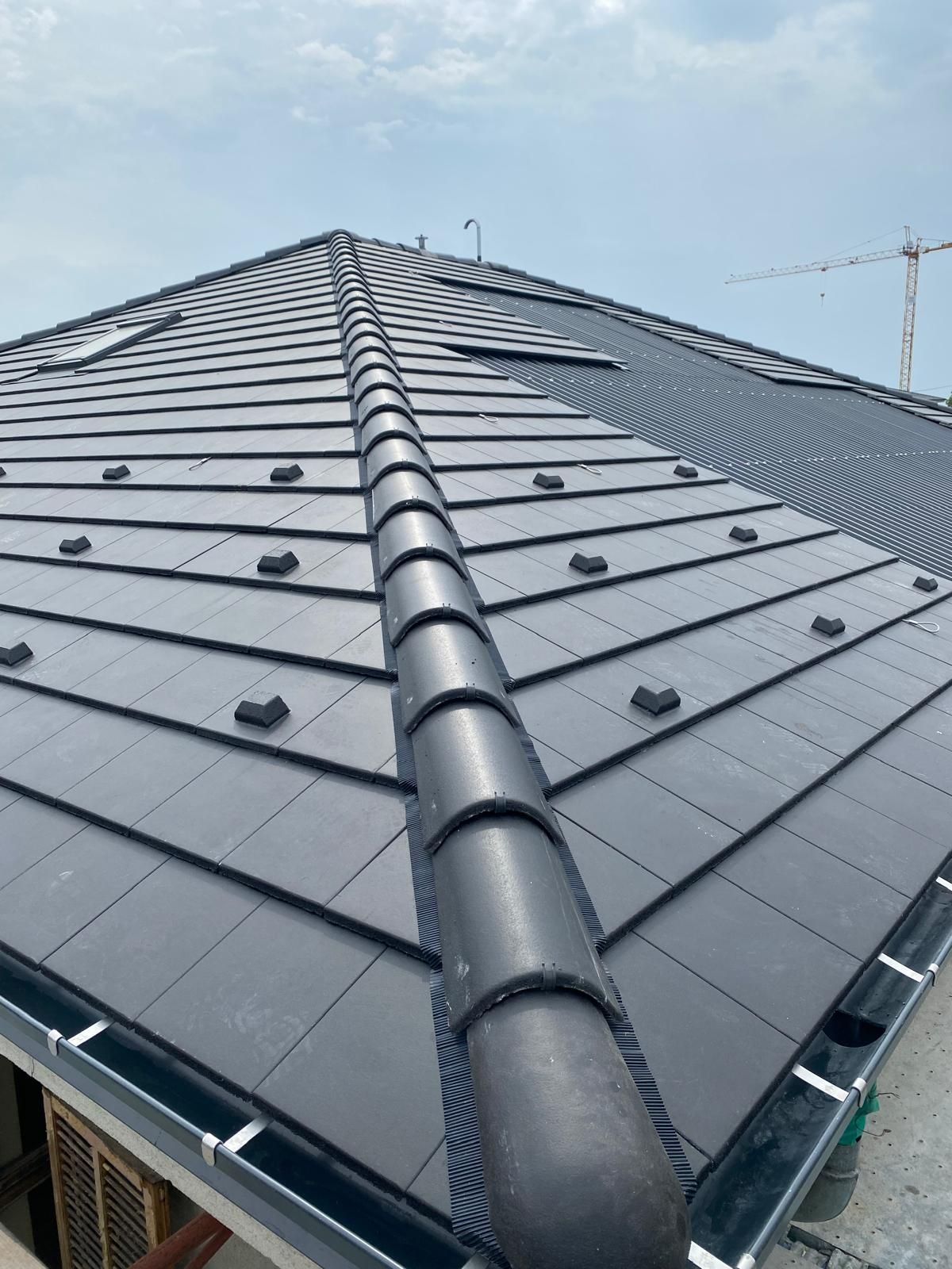 Tetto di tegole grigio scuro con colmo e grondaia neri. Sullo sfondo un cielo azzurro.
