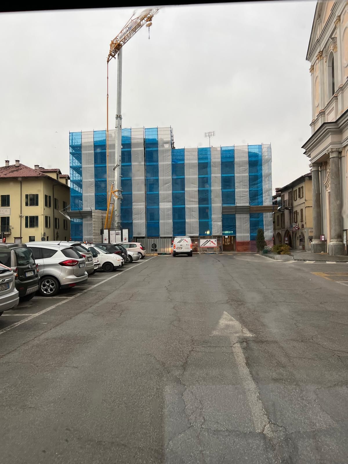 Edificio in costruzione, coperto da reti di sicurezza blu e bianche, in una piazza di una città europea con auto parcheggiate e gru.
