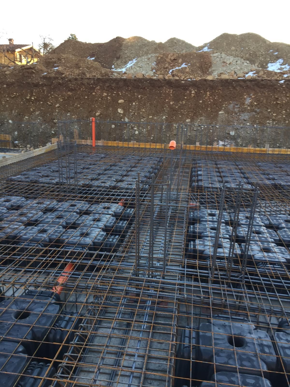 Cantiere con griglia di barre d'armatura e casseforme nere in preparazione per il getto di calcestruzzo. Sullo sfondo un cumulo di terra.