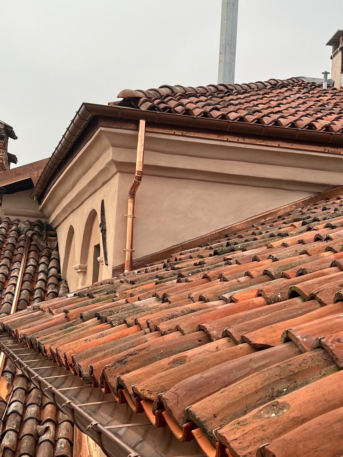 Tetto in tegole rosse con grondaie e pluviali in rame su un edificio beige.