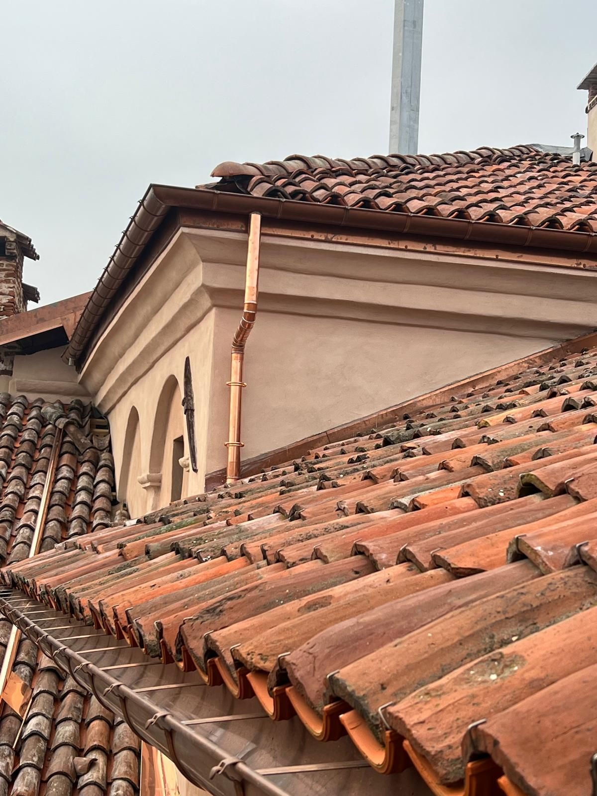 Tetto in tegole rosse con grondaie e pluviale in rame su un edificio con finestre ad arco.