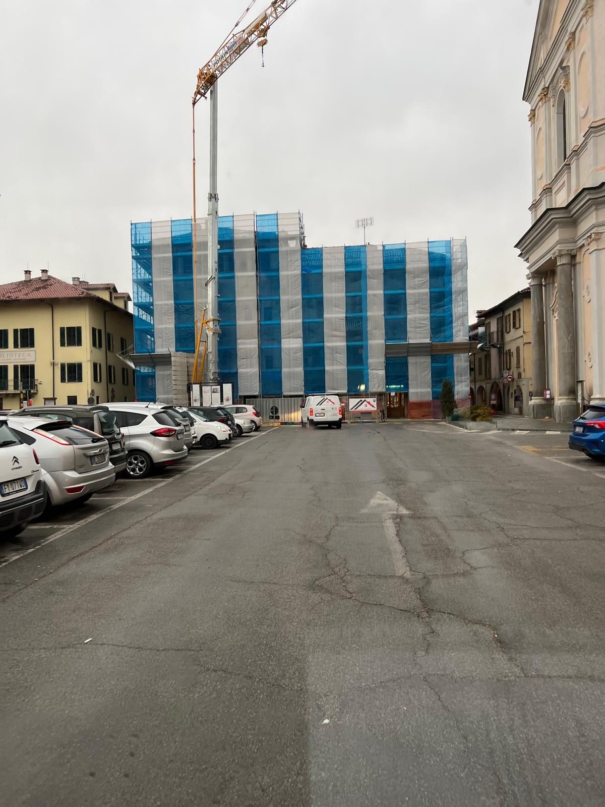 Parcheggio con auto, cantiere coperto da pellicola blu e bianca, gru, cielo nuvoloso.