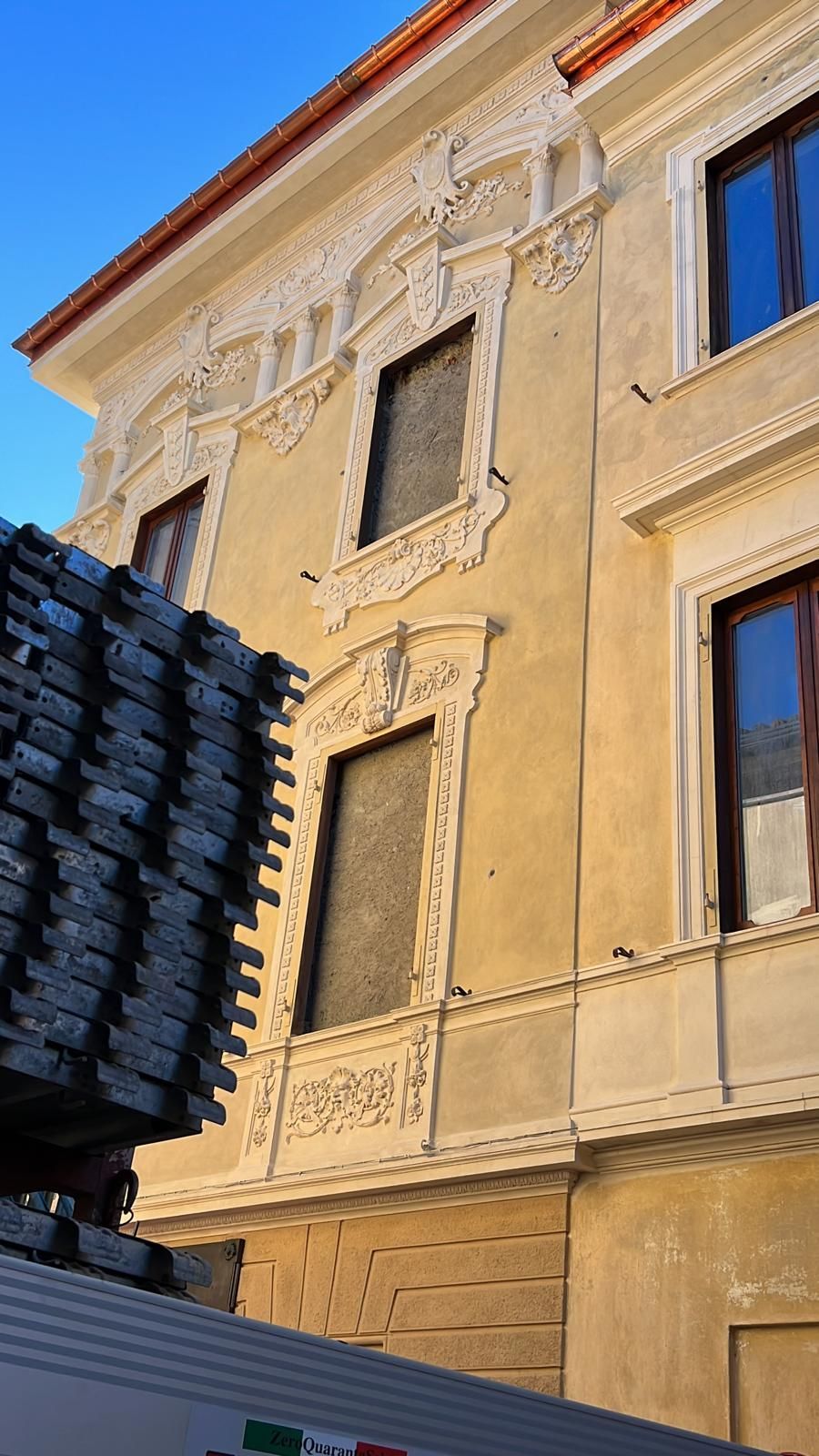 Facciata gialla di un edificio con ornate finiture bianche attorno alle finestre. In primo piano, una pila di materiale edile grigio.