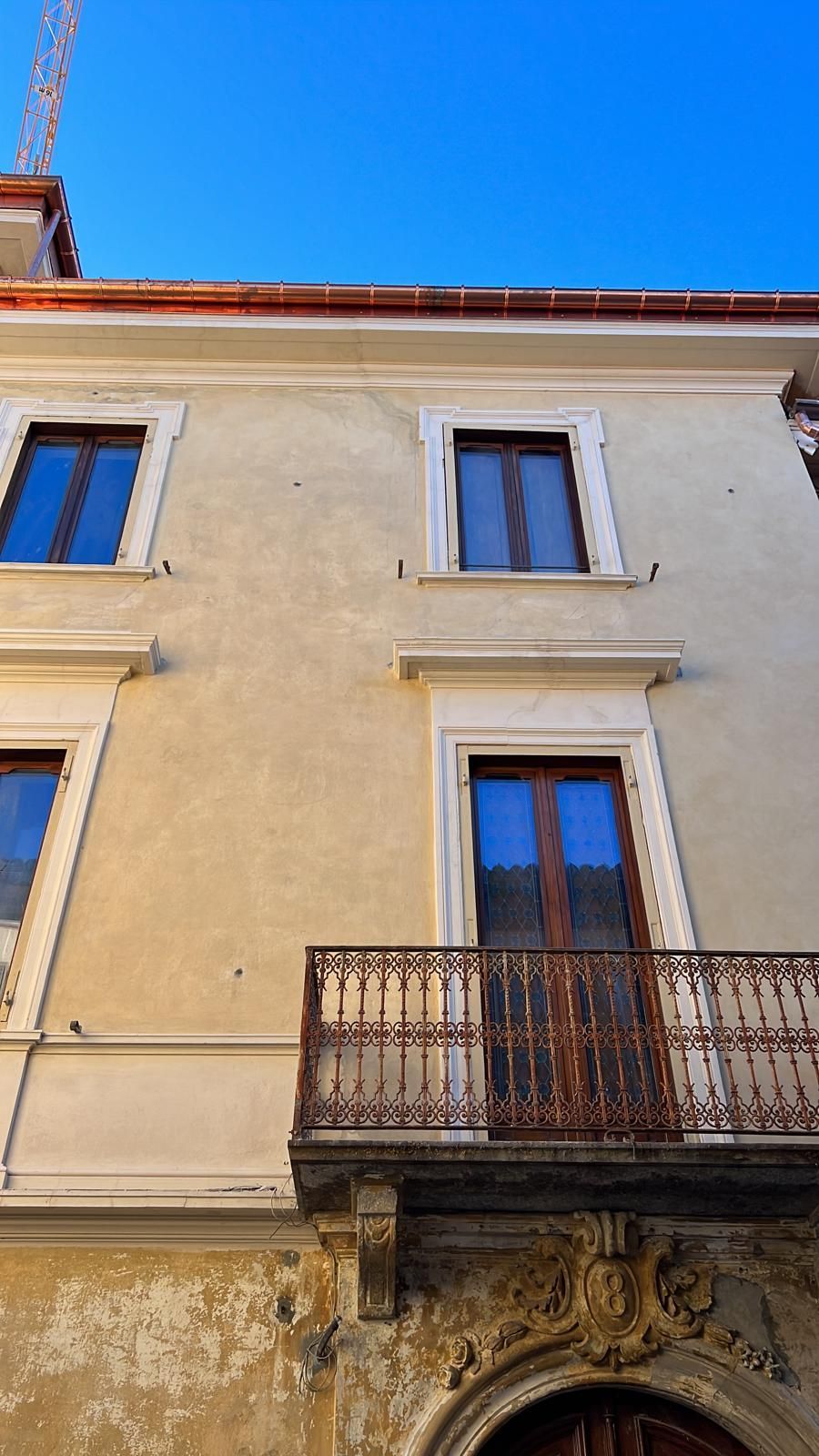 Facciata di un edificio giallo chiaro con finestre, balcone e rifiniture elaborate, contro un cielo azzurro terso.