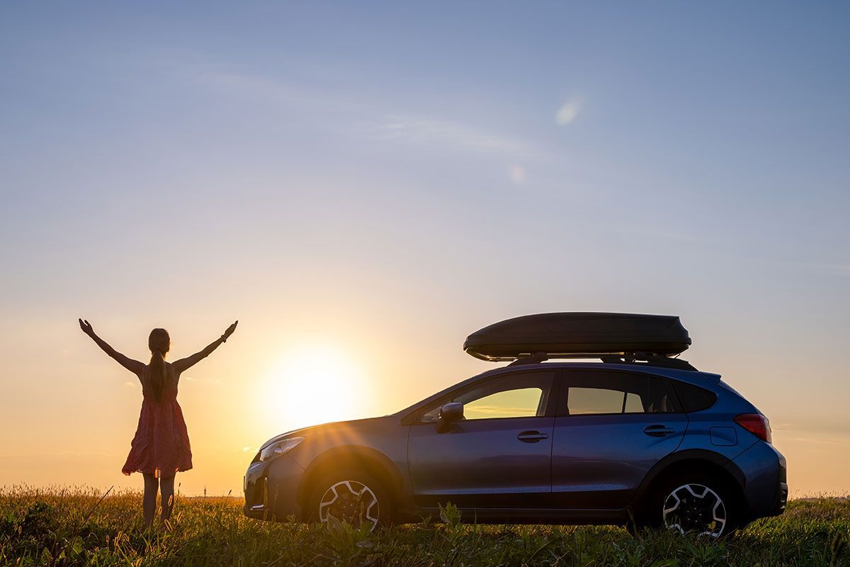 A person stands with arms raised in a field at sunset next to a blue SUV with a cargo carrier on its roof.