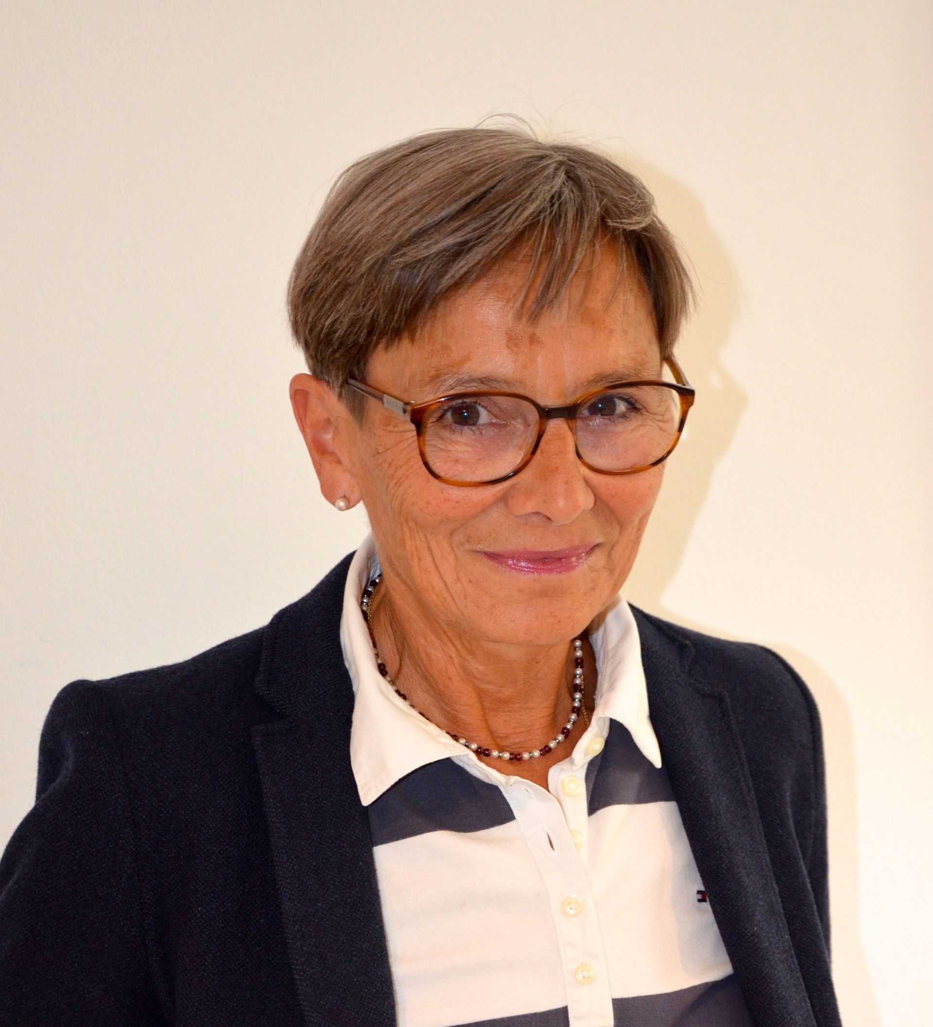 Frau mit kurzem, ergrauendem Haar und Brille, die einen marineblauen Blazer über einem gestreiften Hemd trägt.