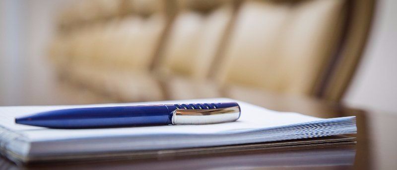 Ein blauer Stift liegt auf einem Notizbuch; im Hintergrund ist unscharf ein Holzstuhl zu sehen.
