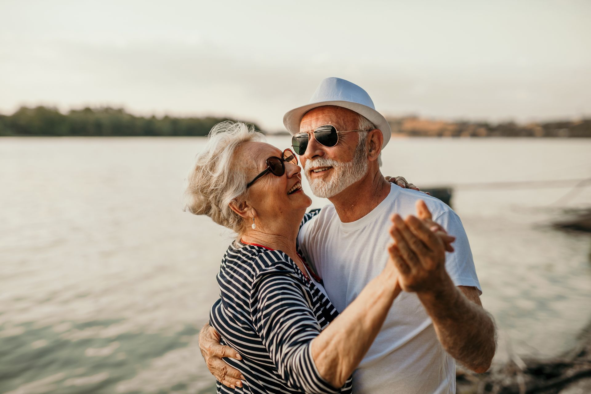 An elderly couple is dancing on the beach near the water.