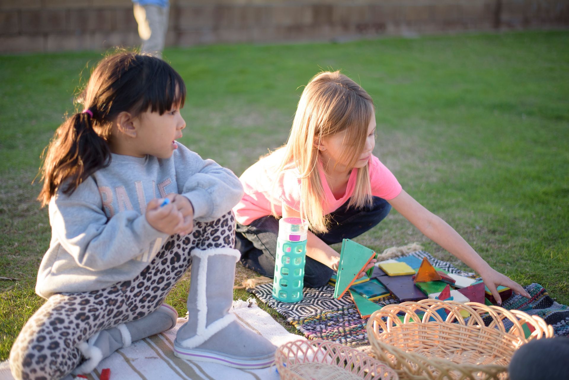 Two montessori children are sitting on a blanket in the grass working with materials.