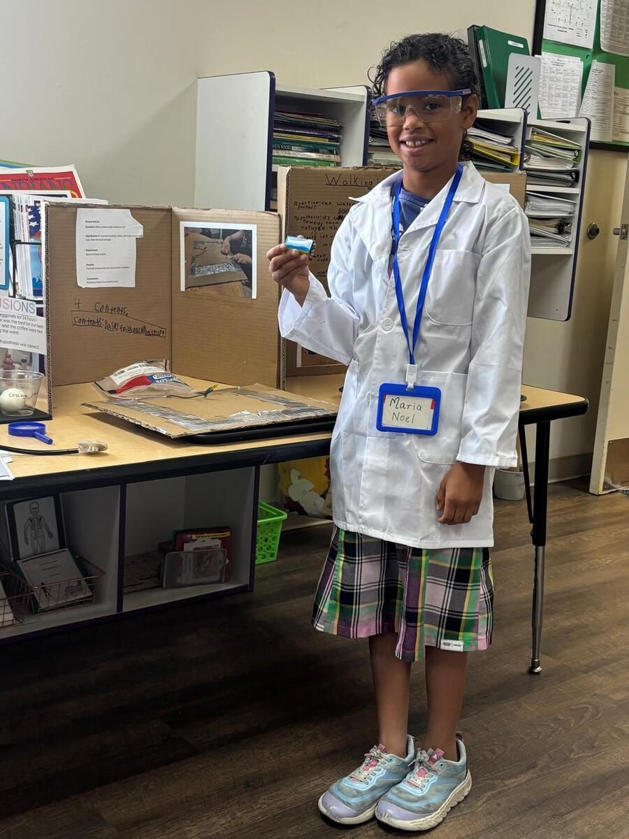 Child in a white lab coat and safety goggles stands by a classroom display, holding a small item.