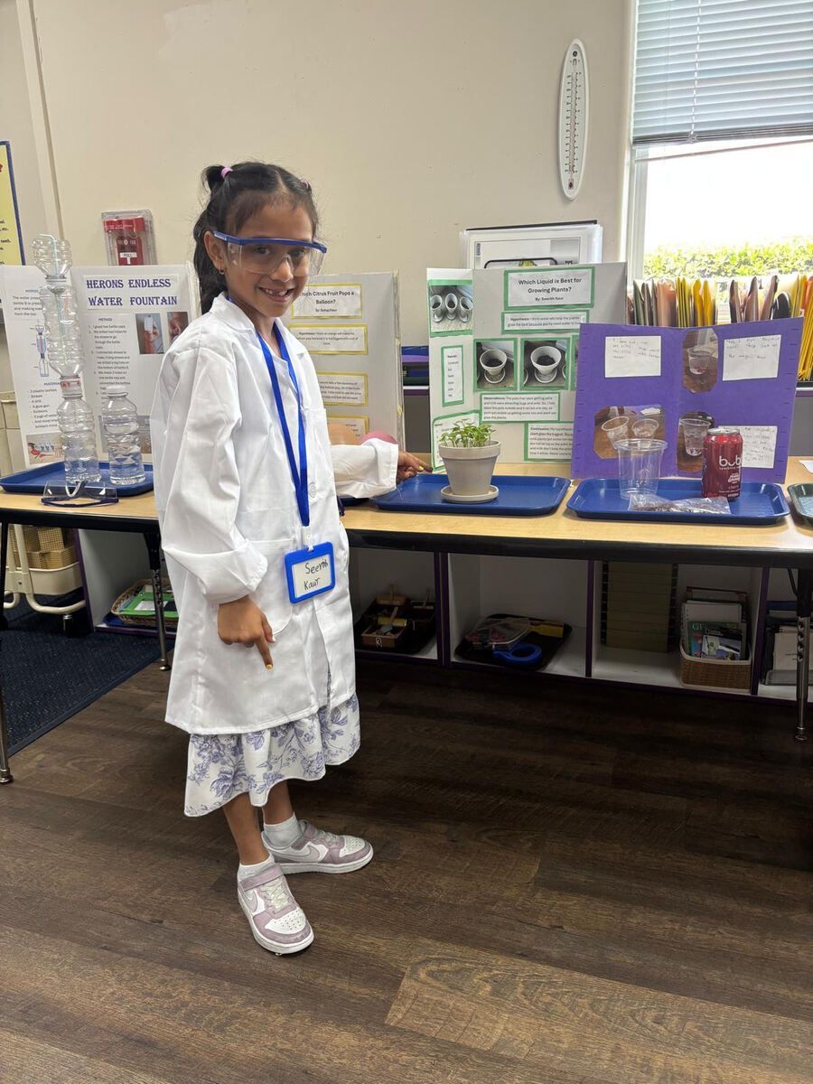 Child in white coat and blue lanyard standing by a science fair display table, smiling at the camera.