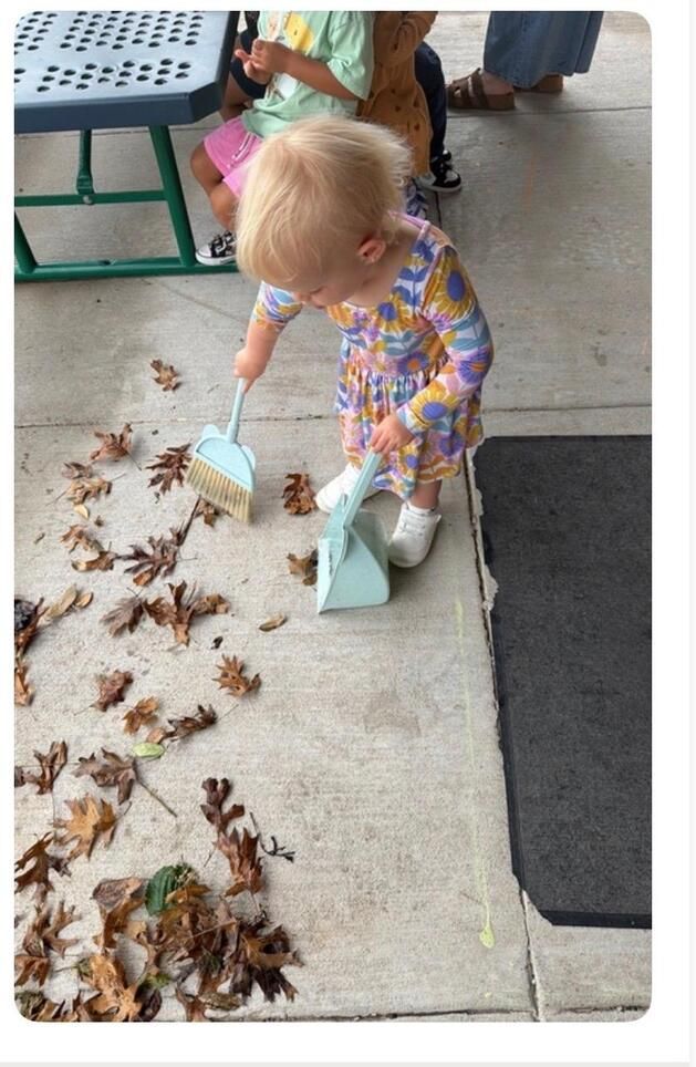 Toddler in colorful outfit sweeping leaves and dirt off a concrete patio with a small broom and dustpan