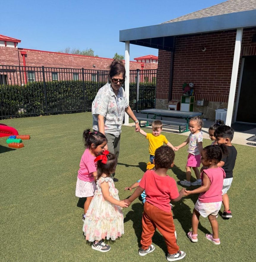 Children holding hands with a teacher in a sunny backyard, standing in a circle on grass near a patio.