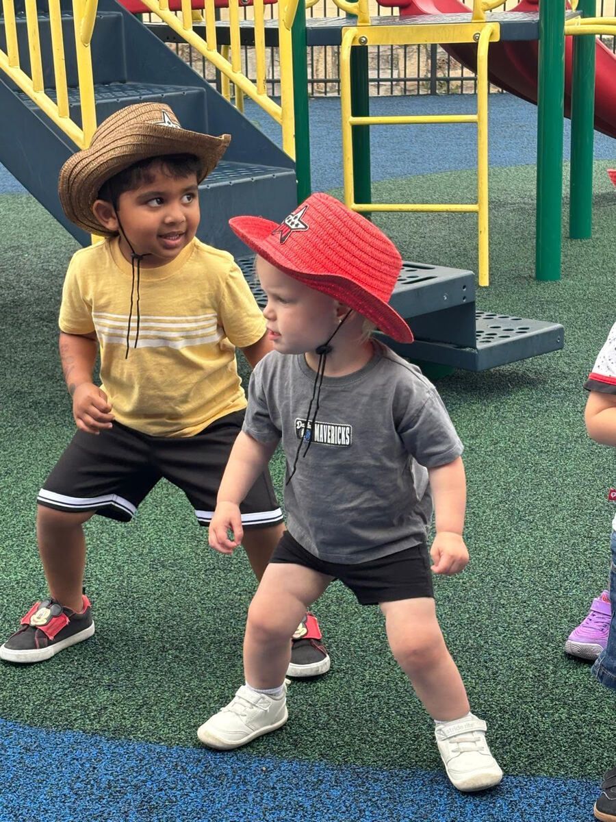 Two toddlers in hats stand on a playground turf, looking at each other near colorful play equipment.